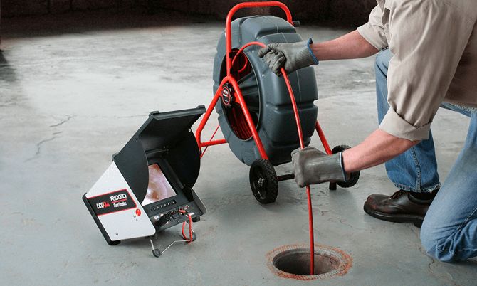 A plumber feeding a drain inspection camera cable from its reel into a floor drain, the camera monitor beside it